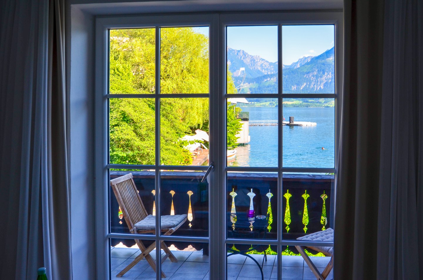 Balkonblick auf einen blauen See mit Schwimmer und Bergen.